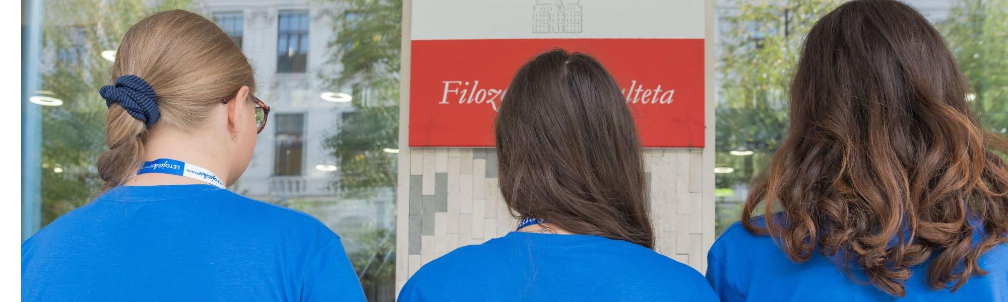 Tri brucke v modrih majicah pred vhodom Filozofske fakultete, gledajo v smeri rdeče table z belim napisom Filozofska fakulteta.